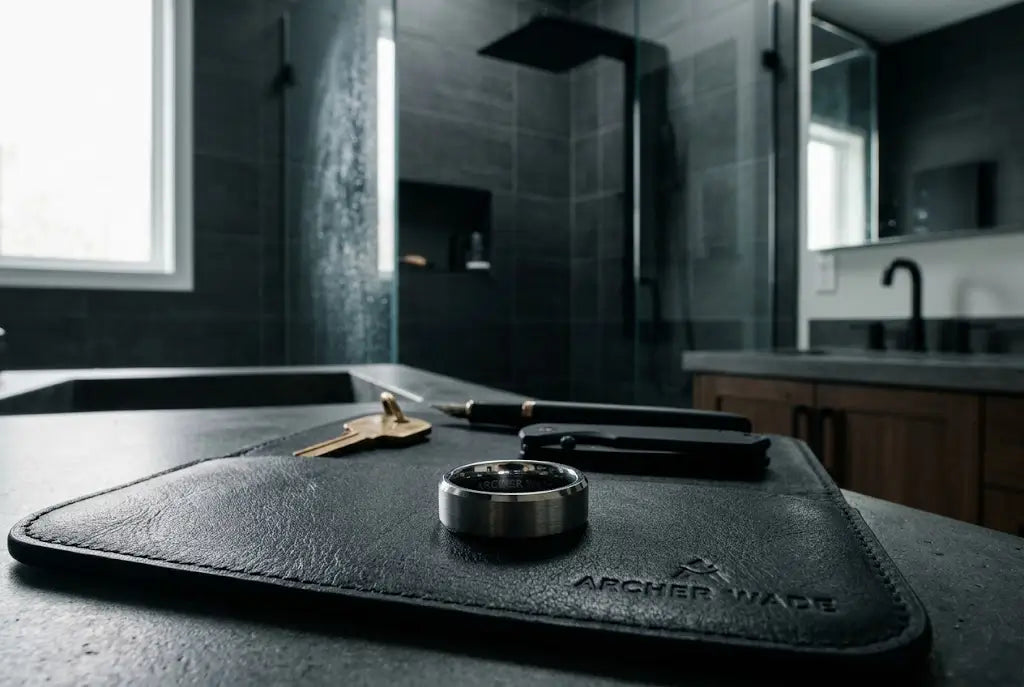 A cinematic, high-quality photograph of a sleek, dark tungsten men's wedding band resting on a premium matte black leather EDC (Everyday Carry) tray.