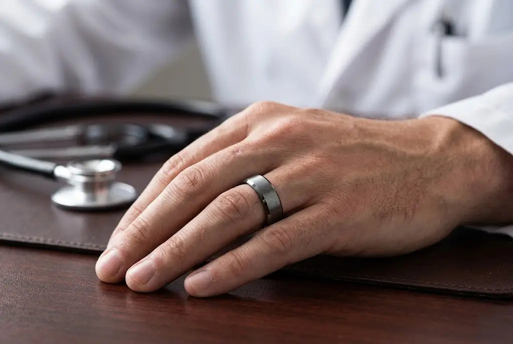 A man's hand wearing a brushed silver tungsten wedding band, with a stethoscope and white coat in soft focus background